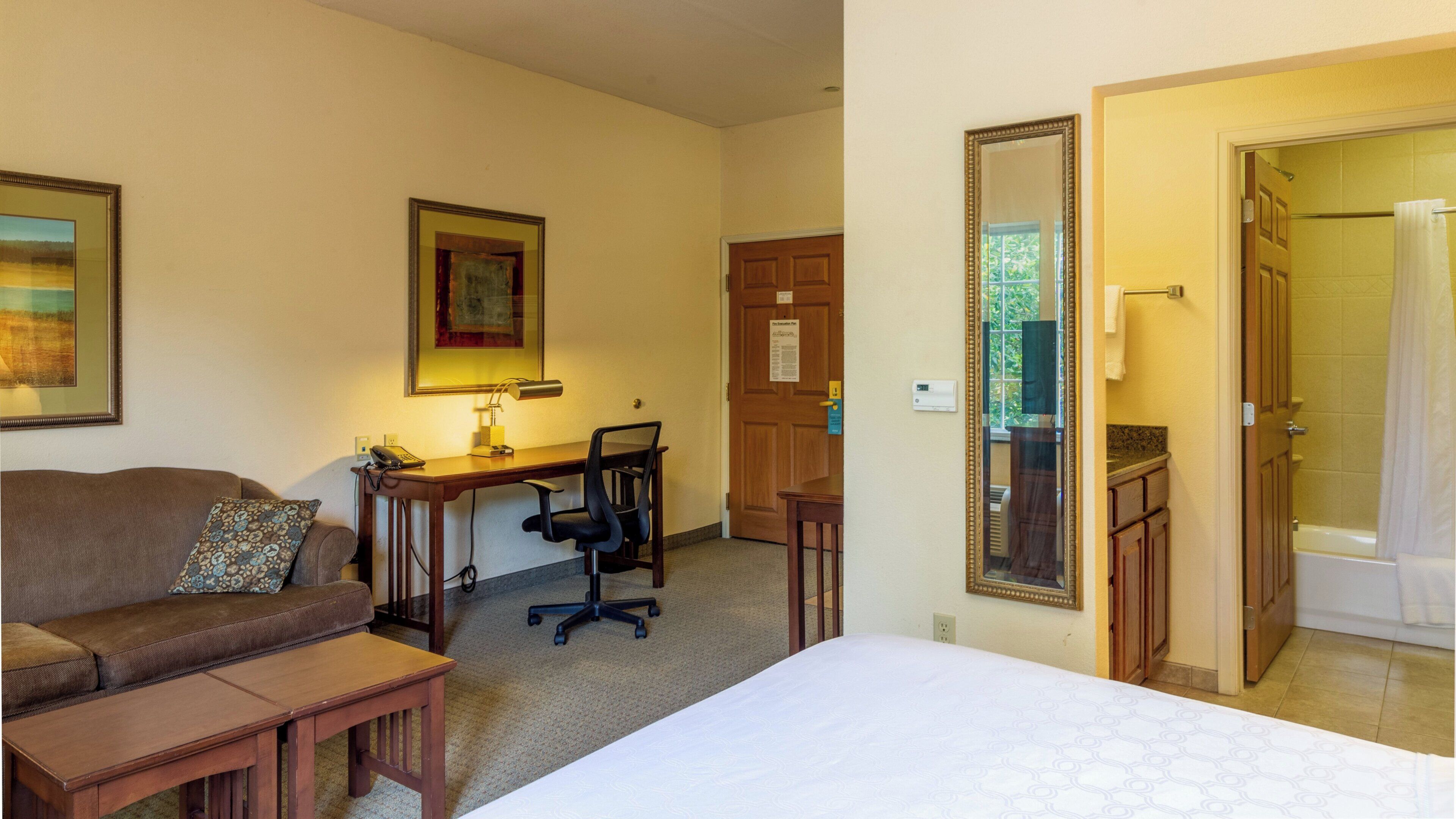 A hotel room with a bed, a desk with a chair, a couch, and a bathroom visible through an open door.