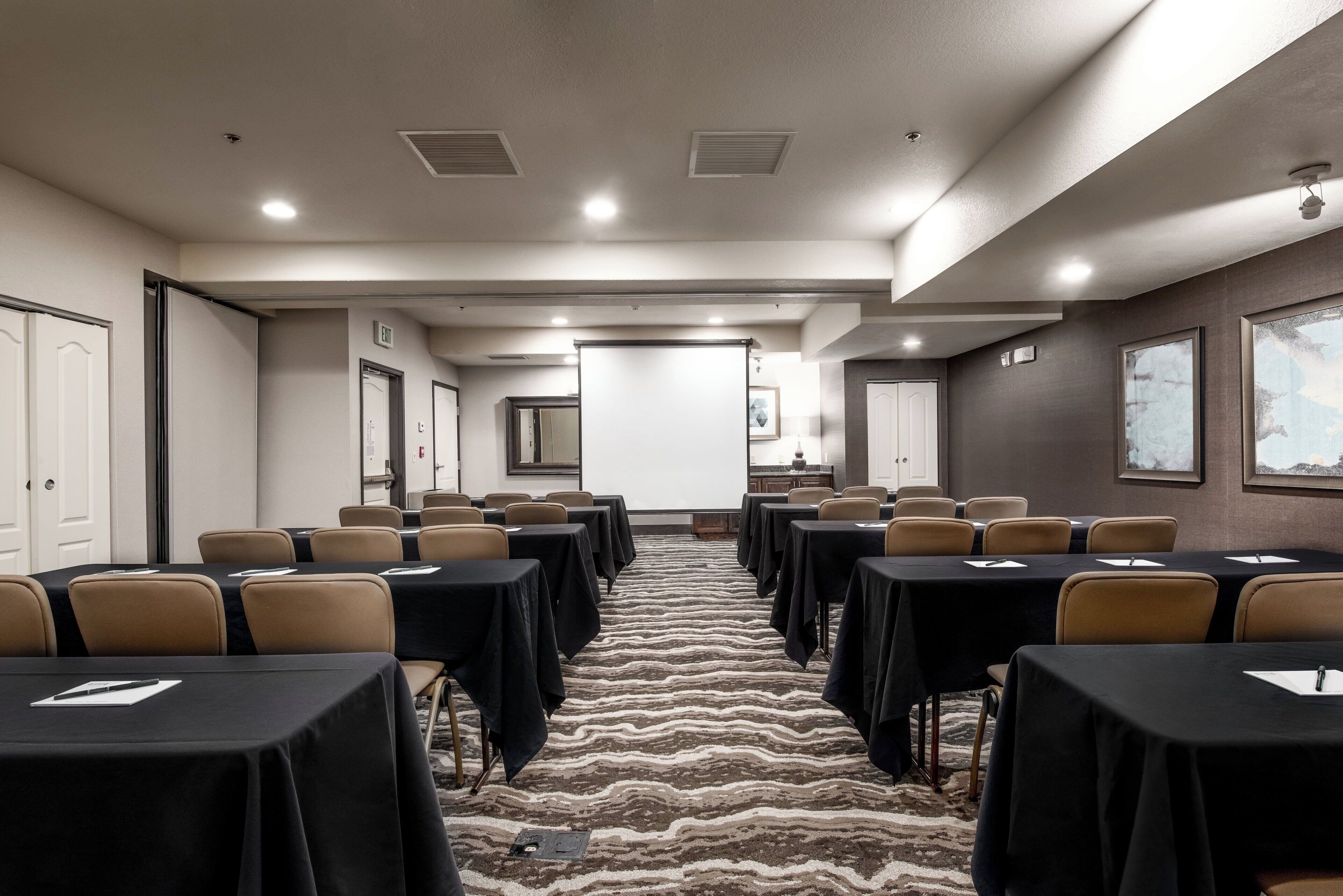 A conference room with rows of tables and chairs, a carpeted floor, and a projector screen at the front.