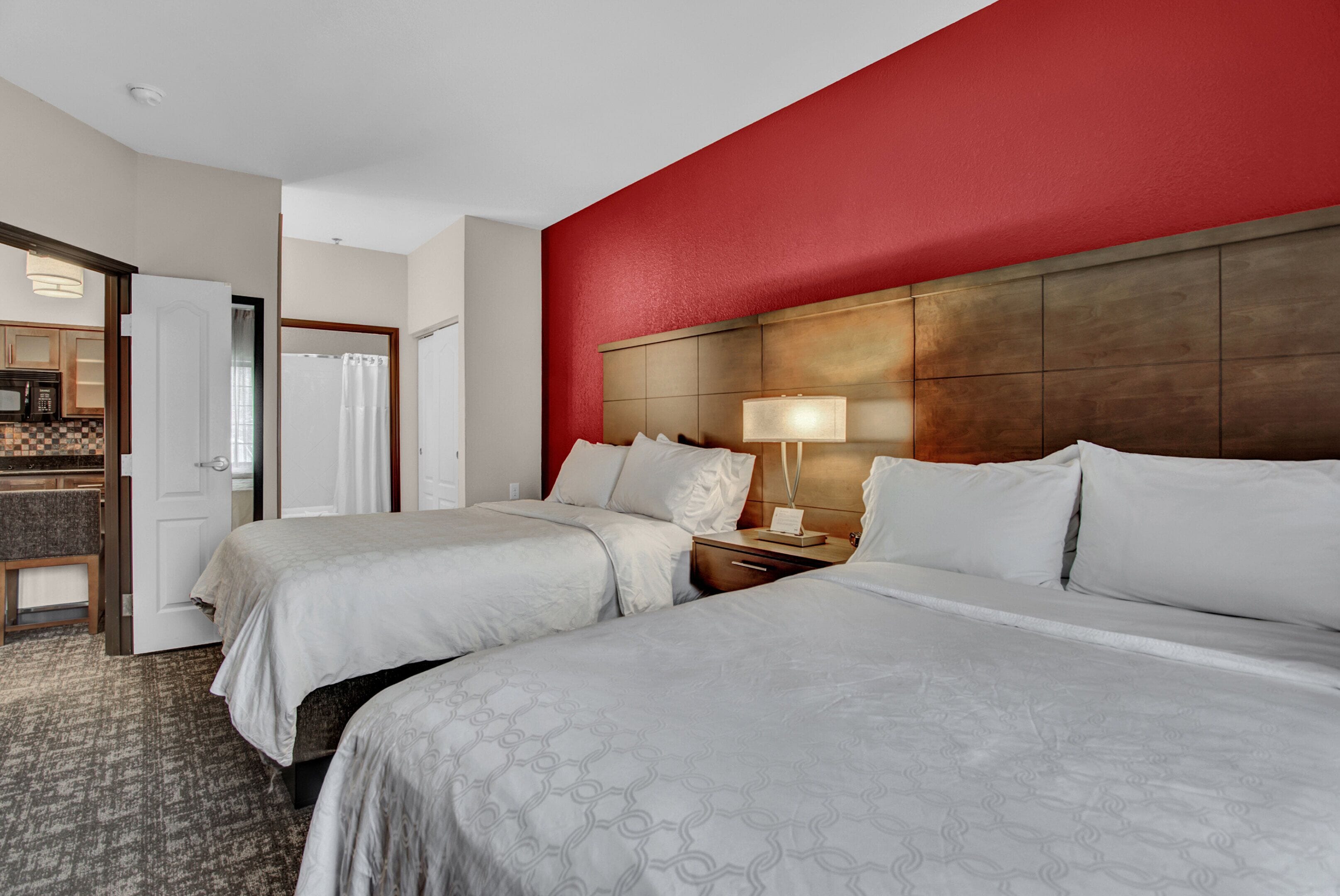 Ein Hotelzimmer mit zwei Betten, einem hölzernen Kopfteil, einem Nachttisch mit Lampe, einem Schreibtisch mit Stuhl und einer Mikrowelle in der Küchenzeile.