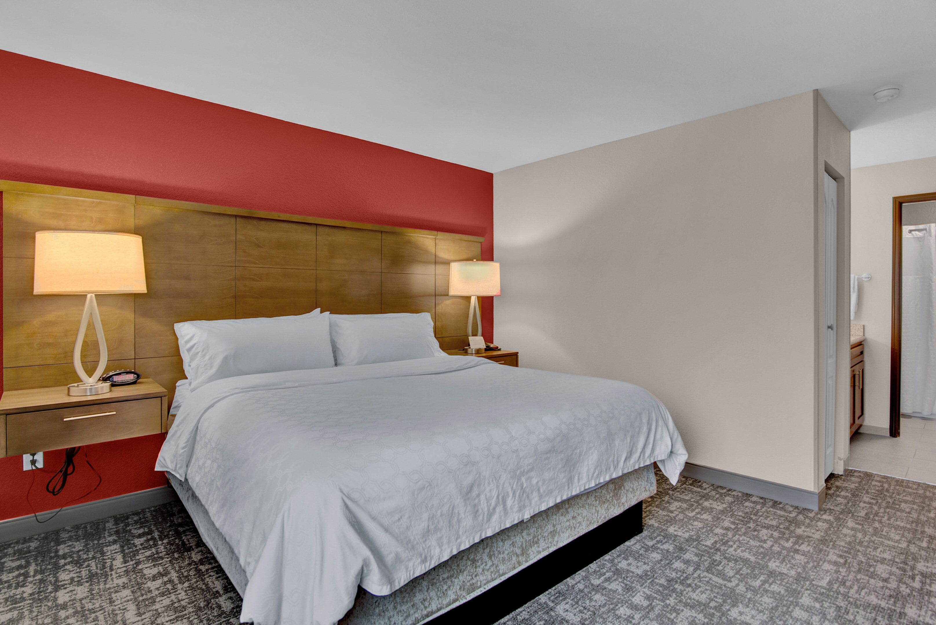 A hotel room with a large bed, two bedside tables with lamps, a red accent wall, and a carpeted floor.