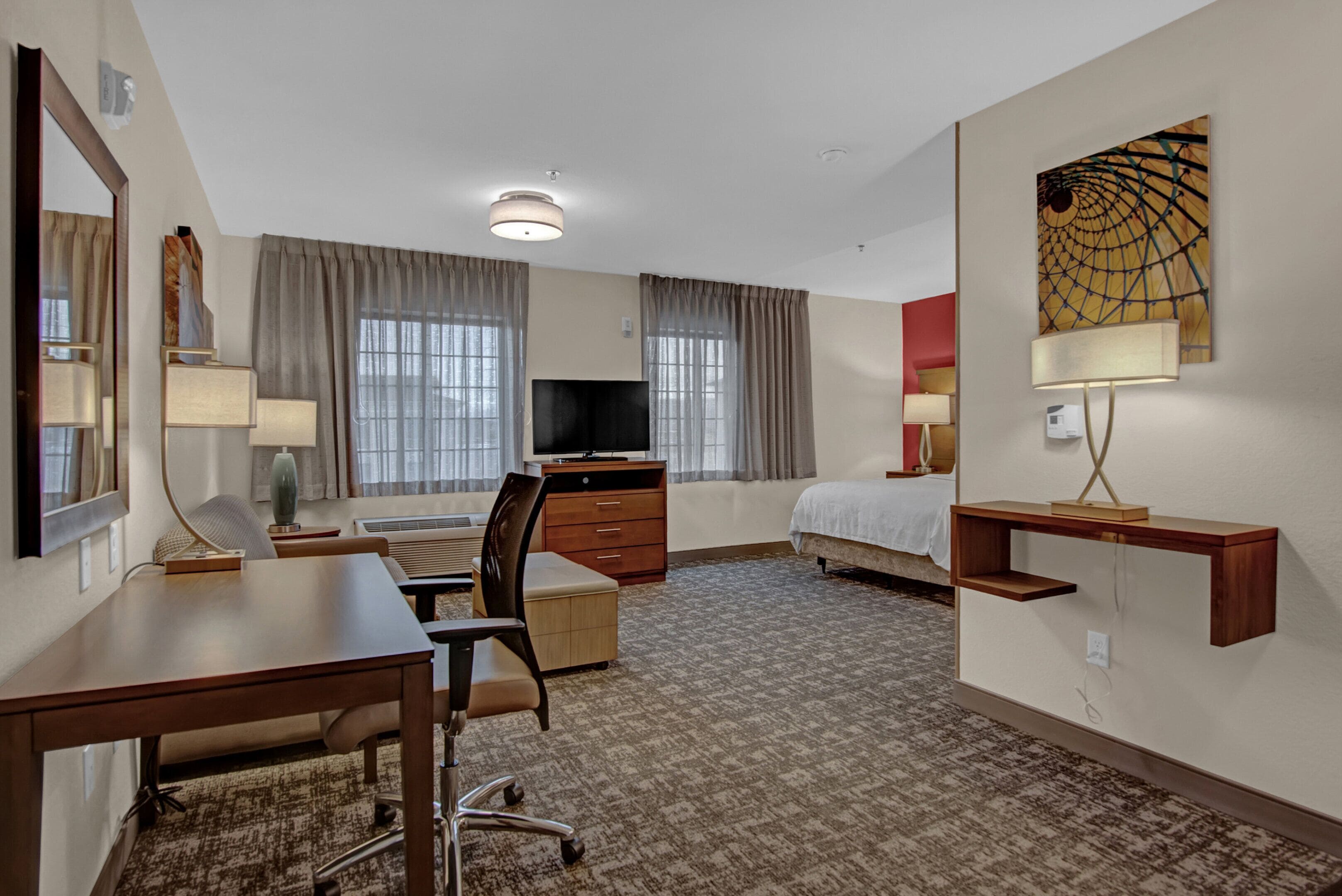 Ett hotellrum med ett skrivbord, en stol, en säng, en TV och ett stort fönster med gardiner.