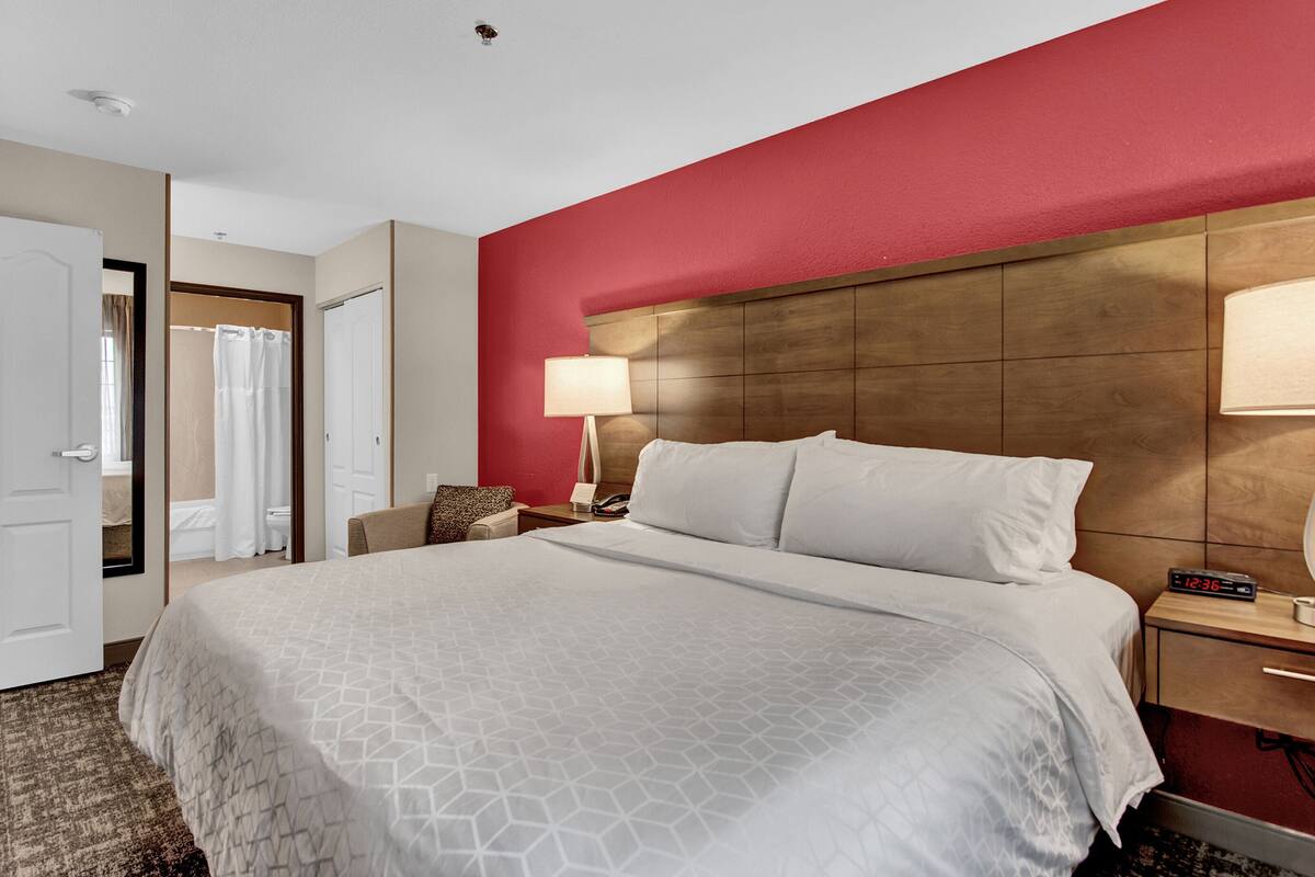 A hotel room with a large bed, a wooden headboard, a bedside table with a lamp, and a red accent wall.