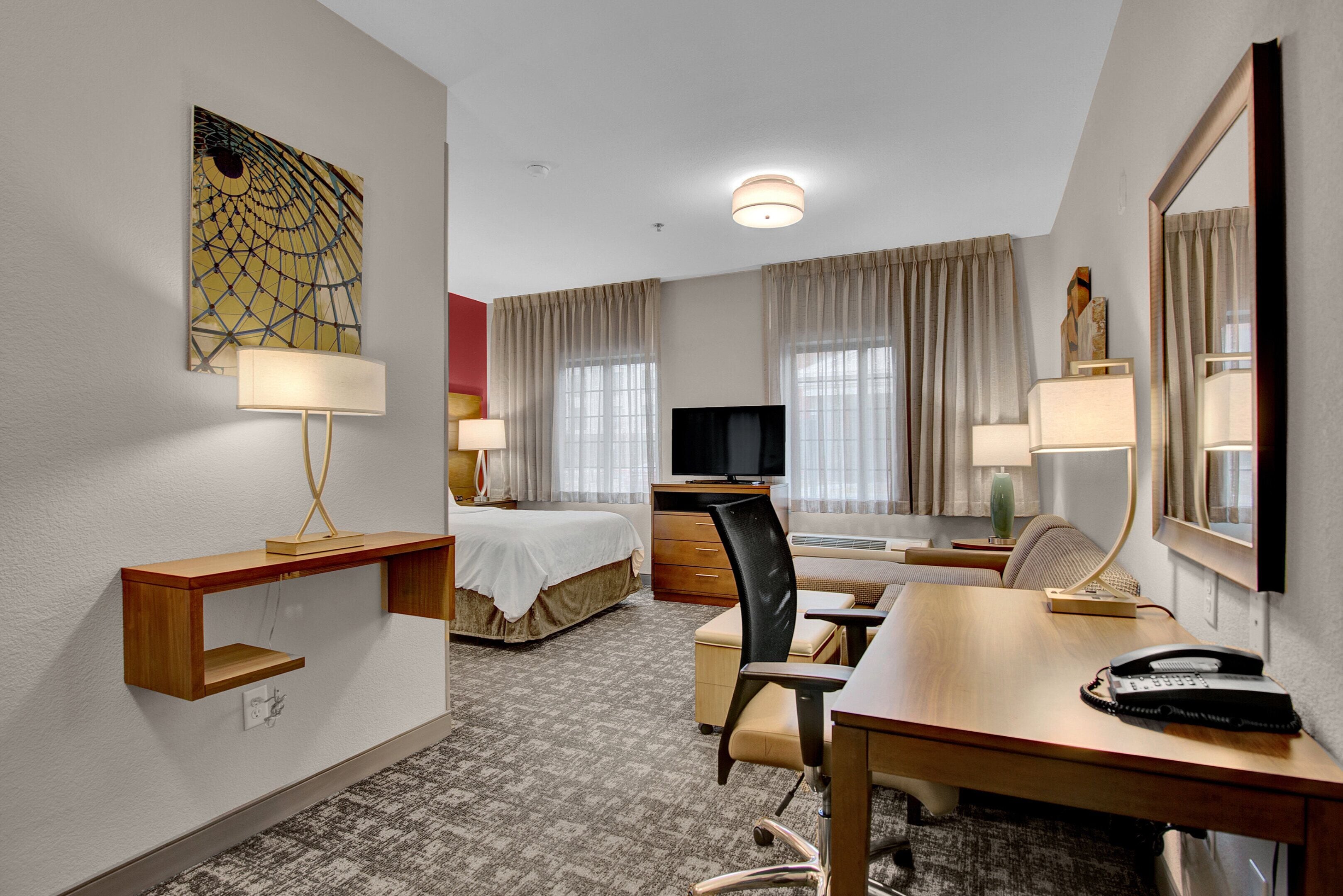 Habitación de hotel con cama, escritorio, silla, televisor y un espejo grande.