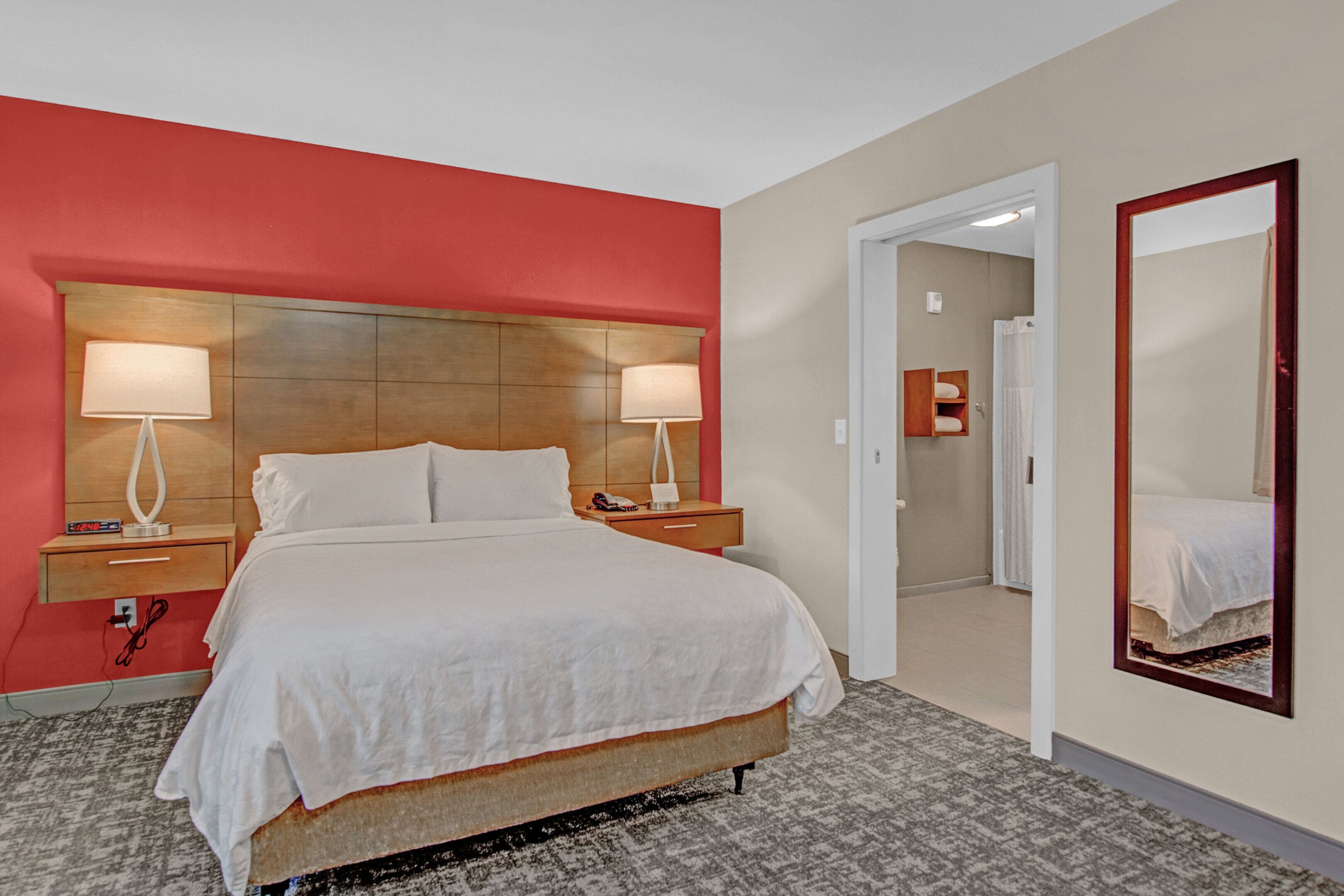 A hotel room with a large bed, two bedside tables with lamps, a wooden headboard, a mirror, and a door leading to a bathroom.