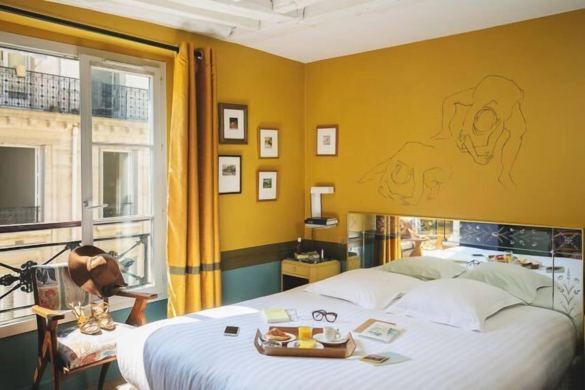 A hotel room with a bed, a desk, a chair, a window with curtains, and framed pictures on the wall.