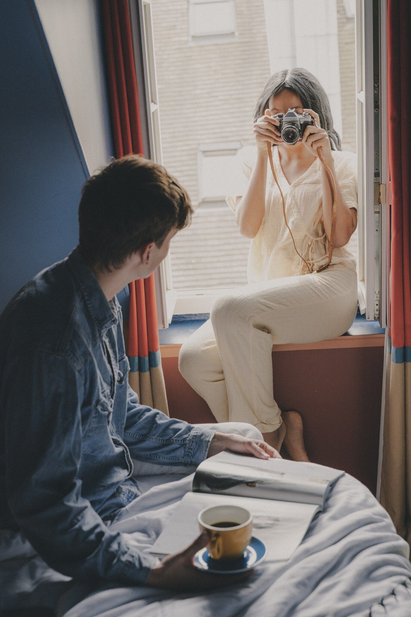 Une personne est assise sur le rebord d’une fenêtre, un appareil photo à la main, et une autre personne est assise sur un lit, un livre et une tasse à ses côtés.
