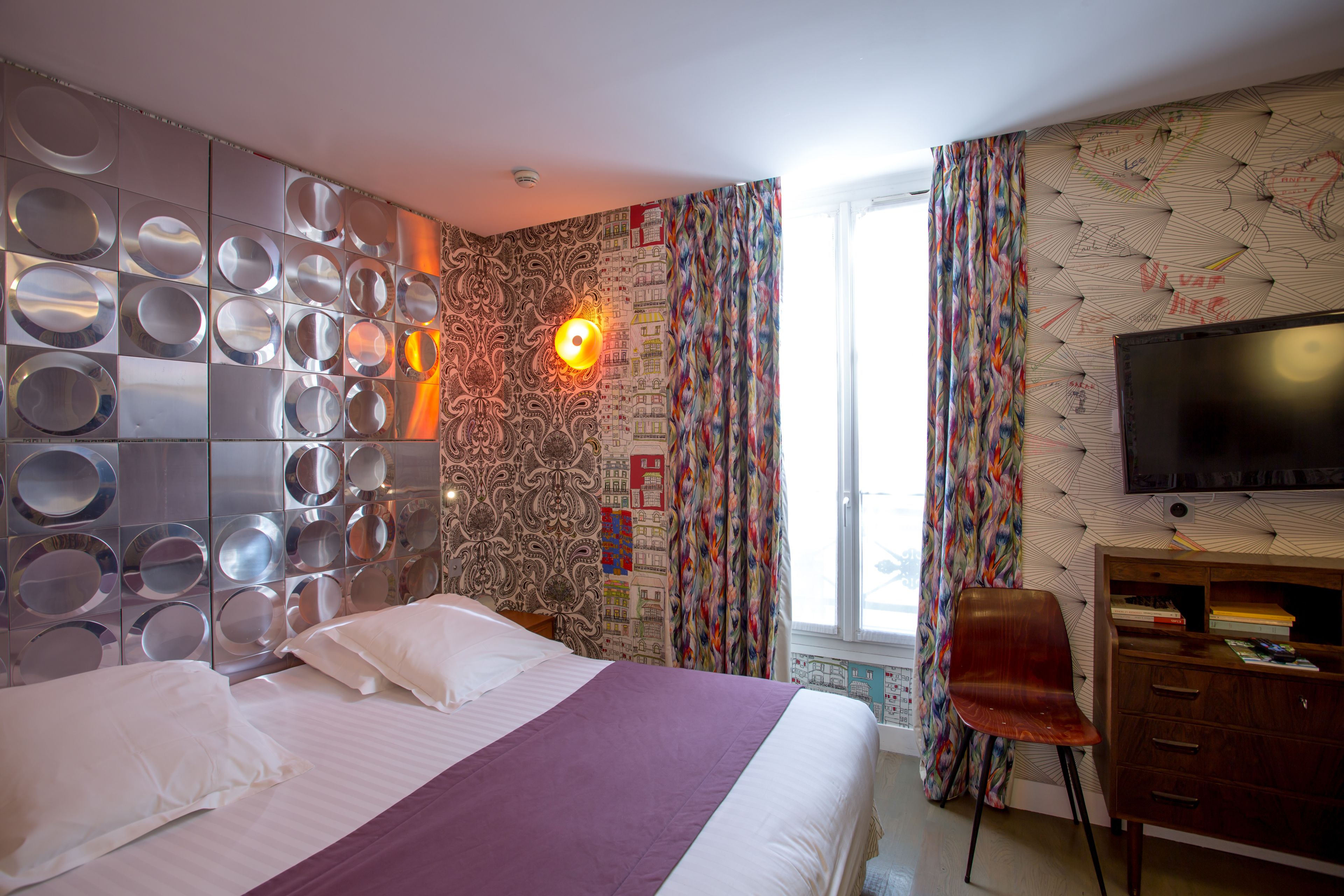 Habitación de hotel con cama, escritorio, silla, televisor y ventana con cortinas.