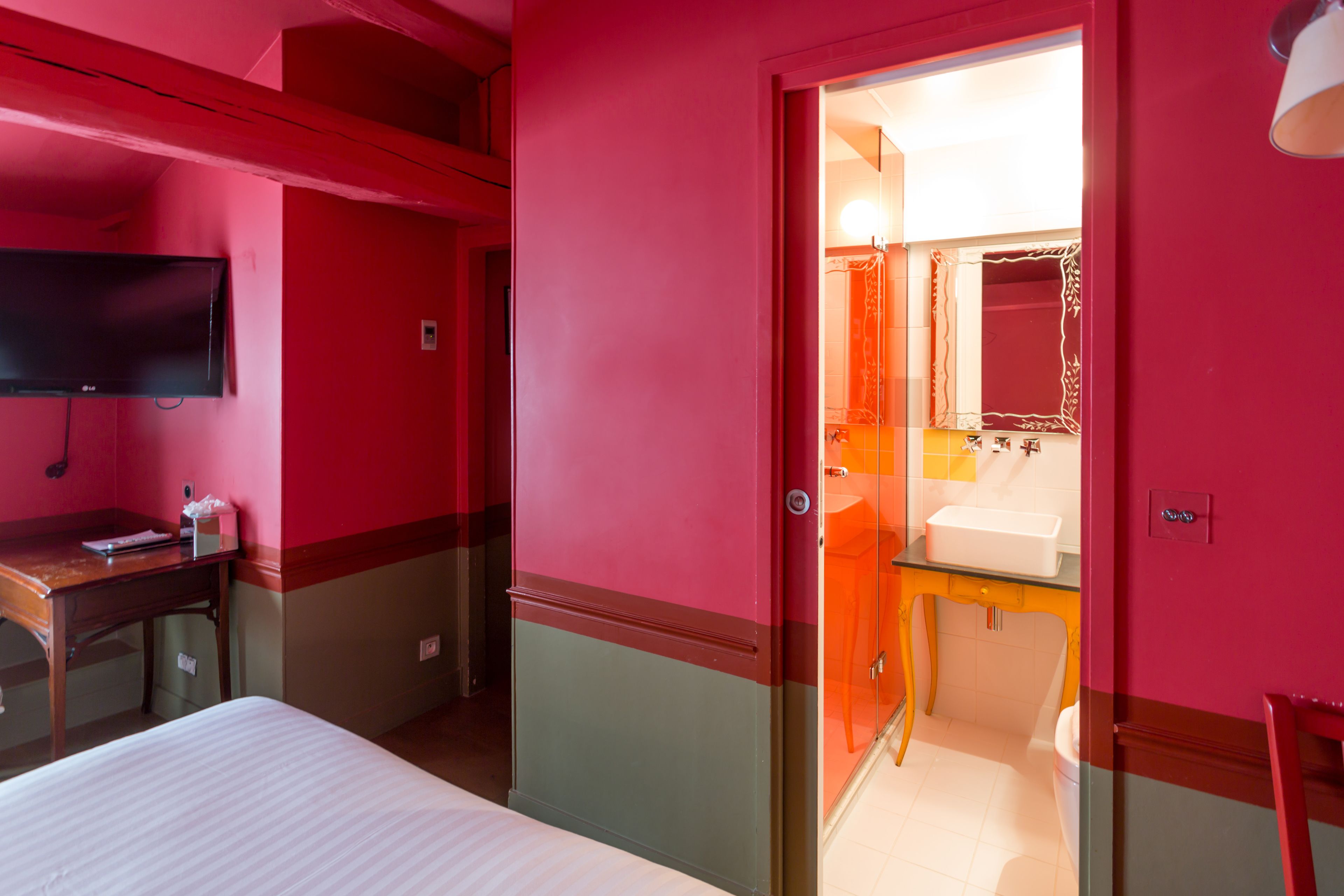 Quarto de hotel com cama, escrivaninha, televisão, banheiro com pia e espelho, e porta que dá para outro cômodo.