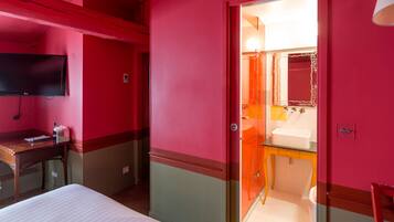 Quarto de hotel com cama, escrivaninha, televisão, banheiro com pia e espelho, e porta que dá para outro cômodo.