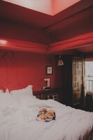 A bedroom with a bed, a bedside table, a lamp, a wall with a line drawing, and a window with curtains.
