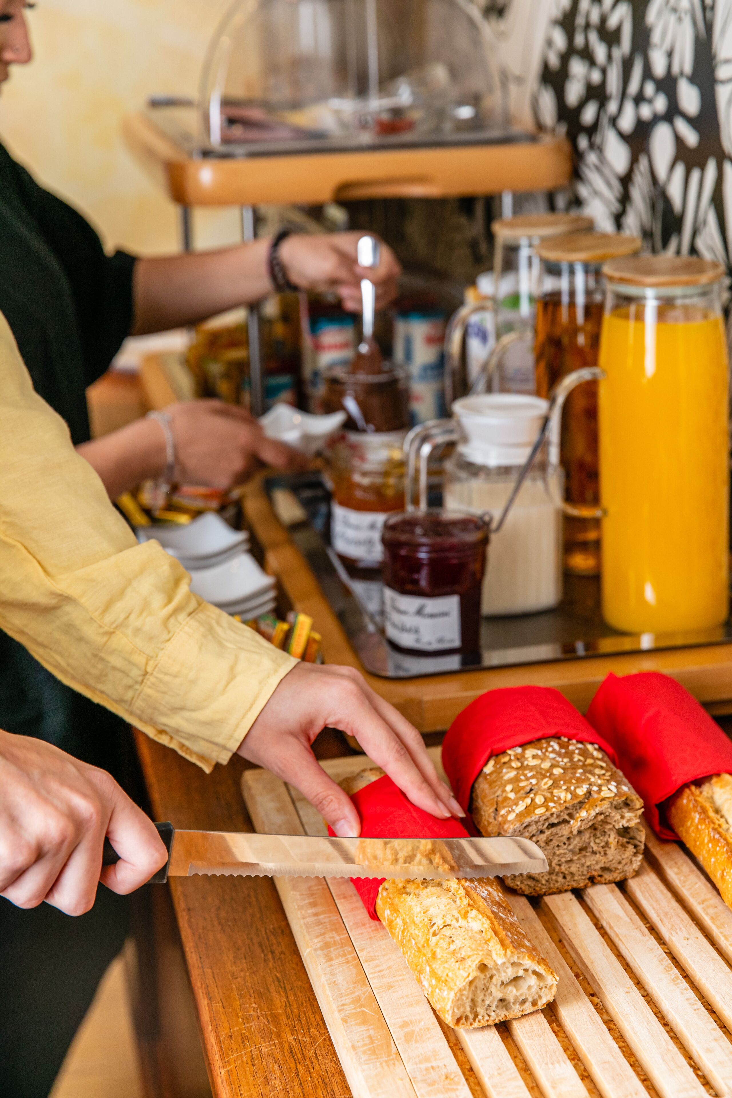 A person slicing bread on a wooden cutting board at a counter with various condiments and a glass container of orange juice.