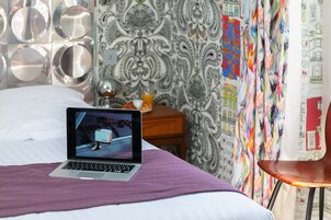 A laptop on a bed with a purple blanket, a wooden nightstand with a lamp and a cup, and a chair.