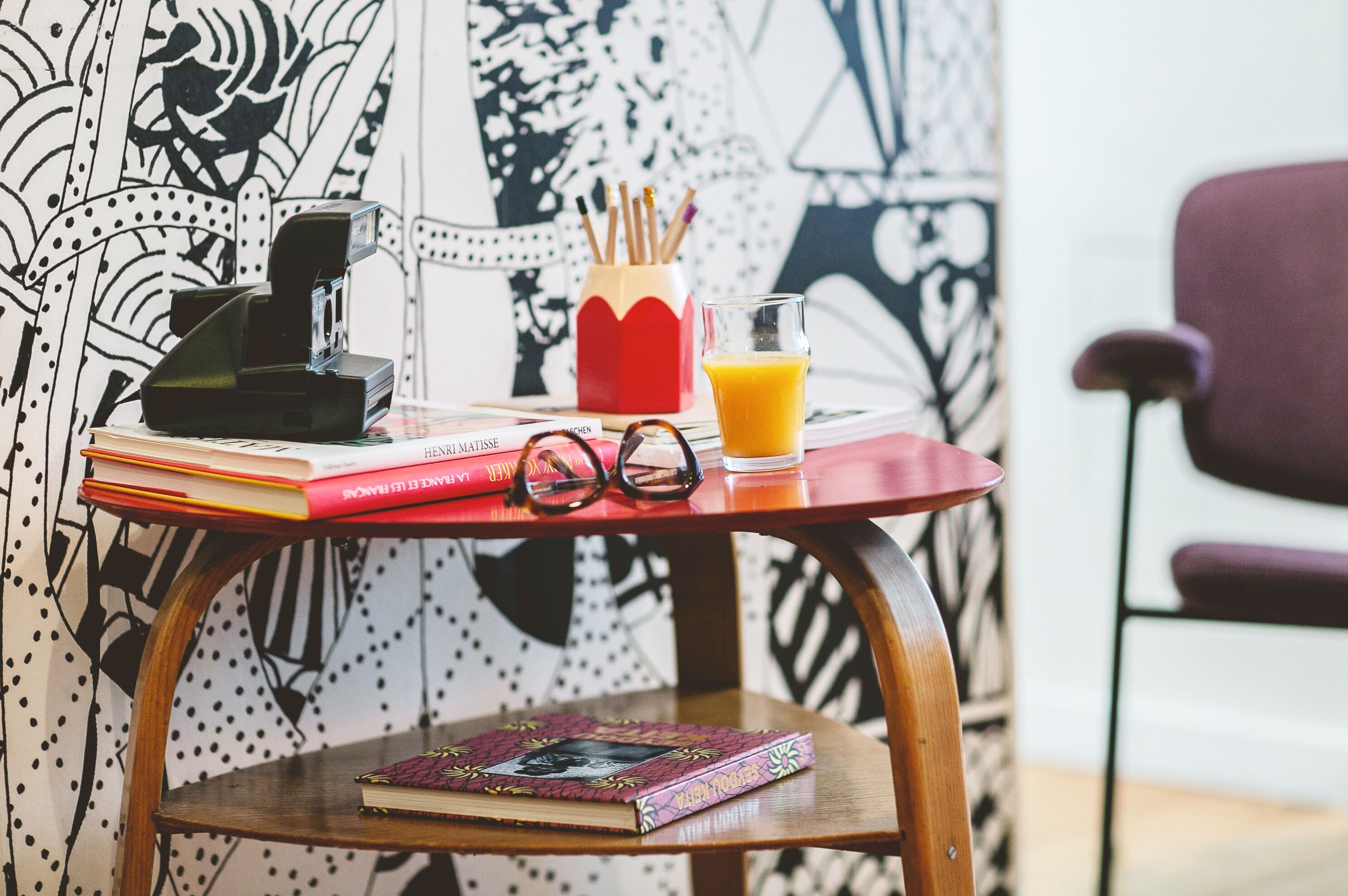 Una mesa con libros, gafas, una cámara y un vaso de jugo de naranja.