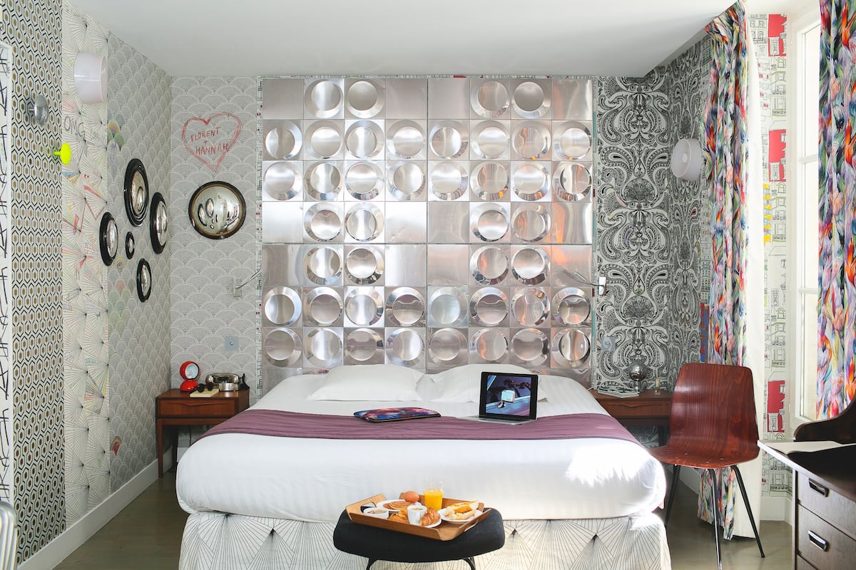 A hotel room with a large bed, a bedside table with a lamp, a chair, and a desk with a computer.