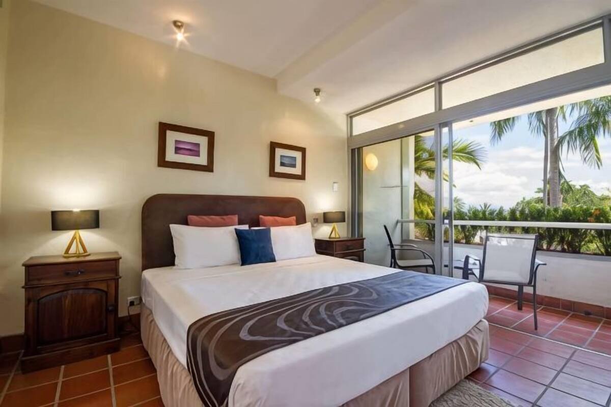 A hotel room with a large bed, two bedside tables, a balcony with a chair, and a view of palm trees.