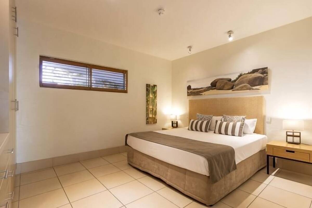 A bedroom with a bed, bedside tables, a window with blinds, and a headboard with a painting.