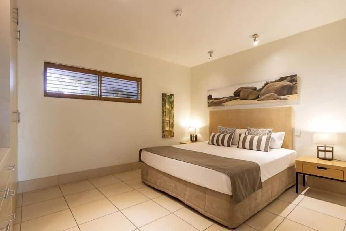 A bedroom with a bed, bedside tables, a window with blinds, and a headboard with a painting.