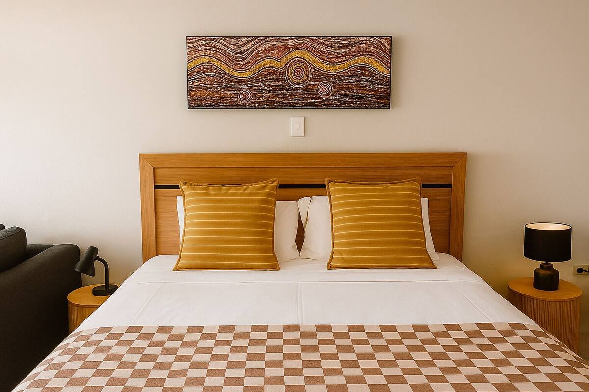 A hotel room with a wooden headboard, two pillows, a checkered bedspread, a lamp on a bedside table, and a wall art piece.