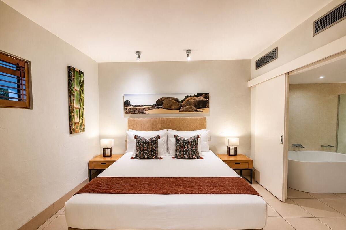 A hotel room with a large bed, two bedside tables with lamps, a painting above the bed, a window with blinds, and a bathroom with a bathtub.
