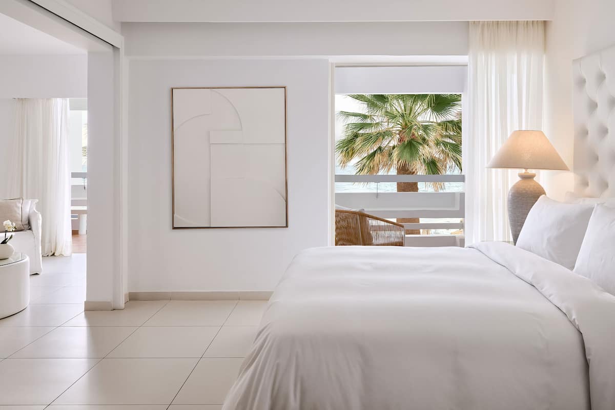 A modern hotel room with a large bed, a bedside lamp, a framed abstract artwork, a view of palm trees, and a balcony with wicker furniture.