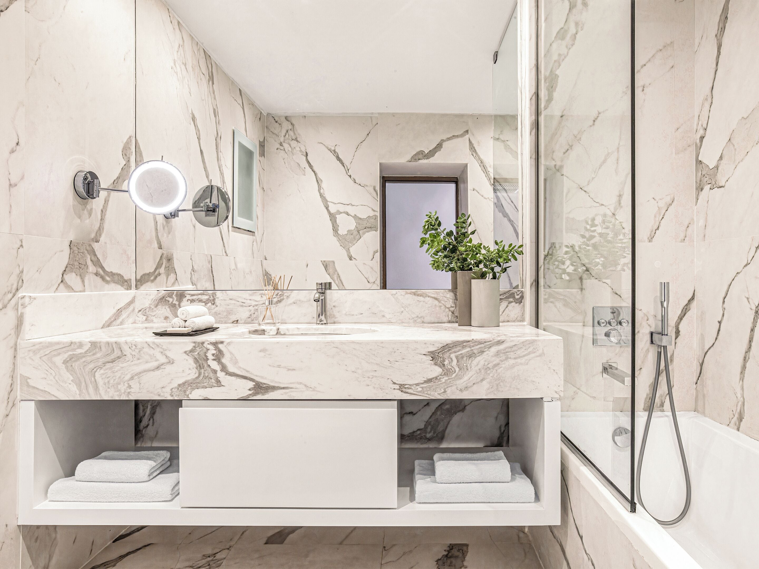 A modern bathroom with marble walls, a glass shower enclosure, and a built-in vanity with a mirror and towel storage.
