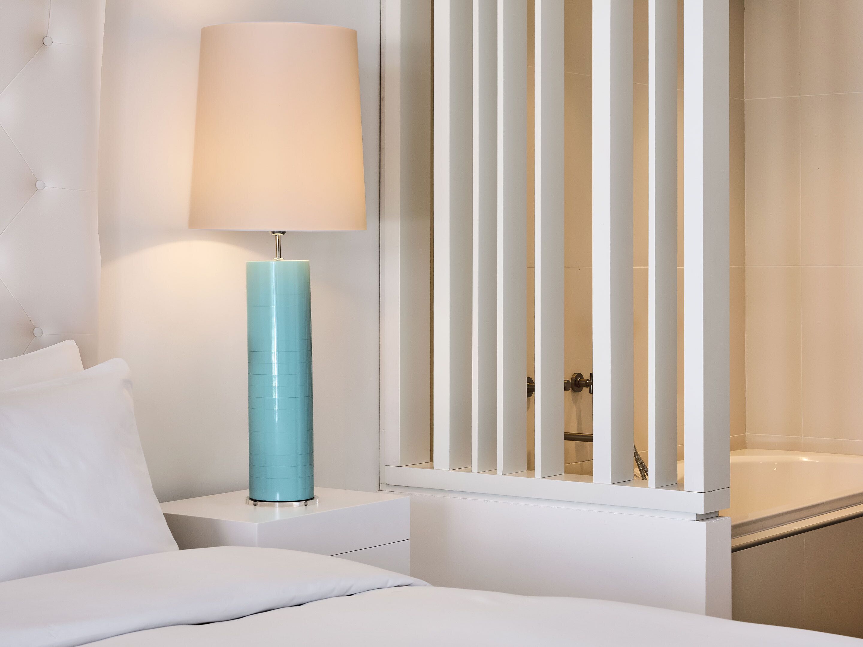 Un dormitorio moderno con una cama, una mesita de noche blanca, una lámpara azul y vistas a un baño con bañera.