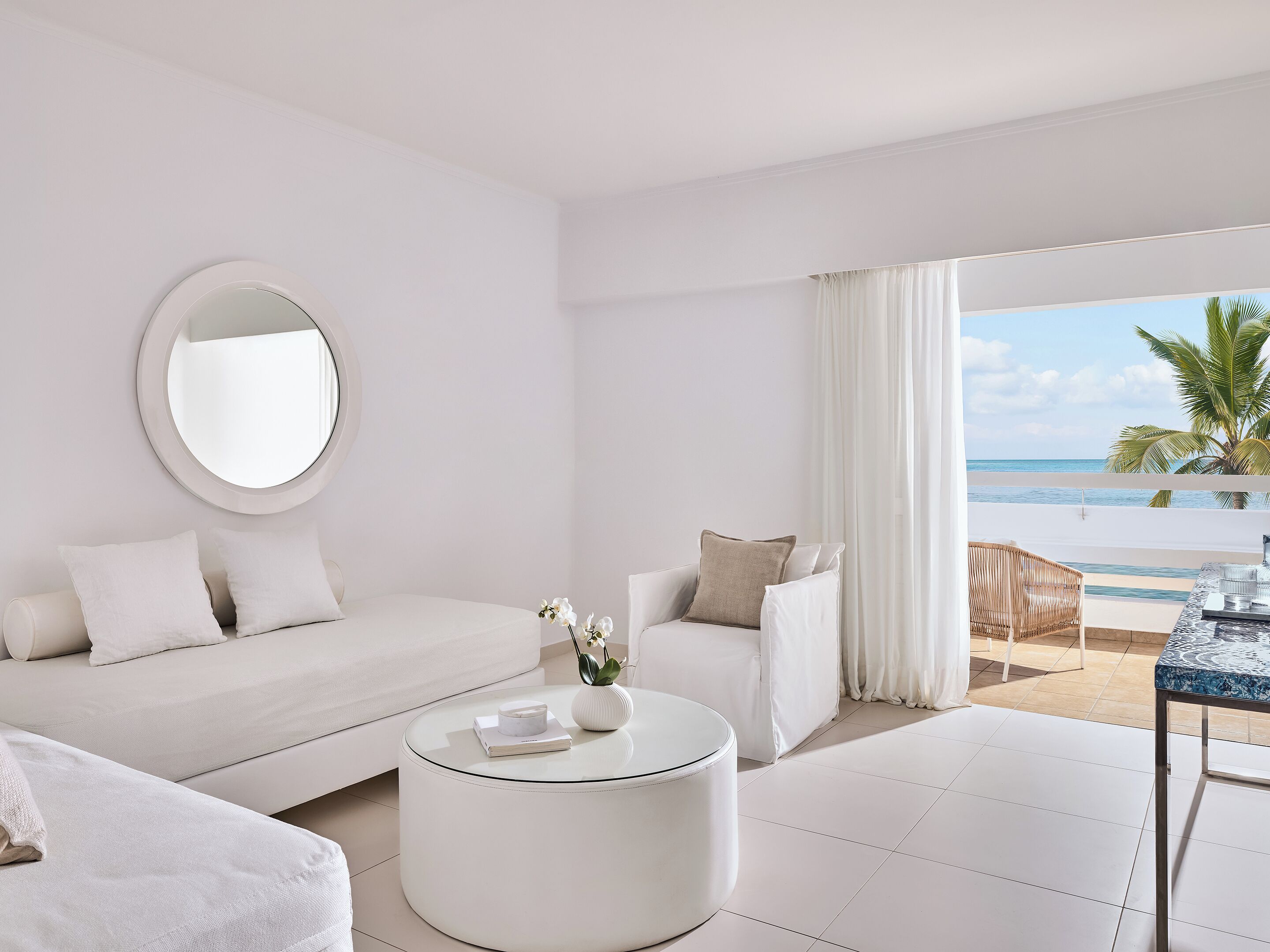 Une chambre d’hôtel moderne aux couleurs claires, avec un miroir rond, un canapé, une table basse et une vue sur la plage.