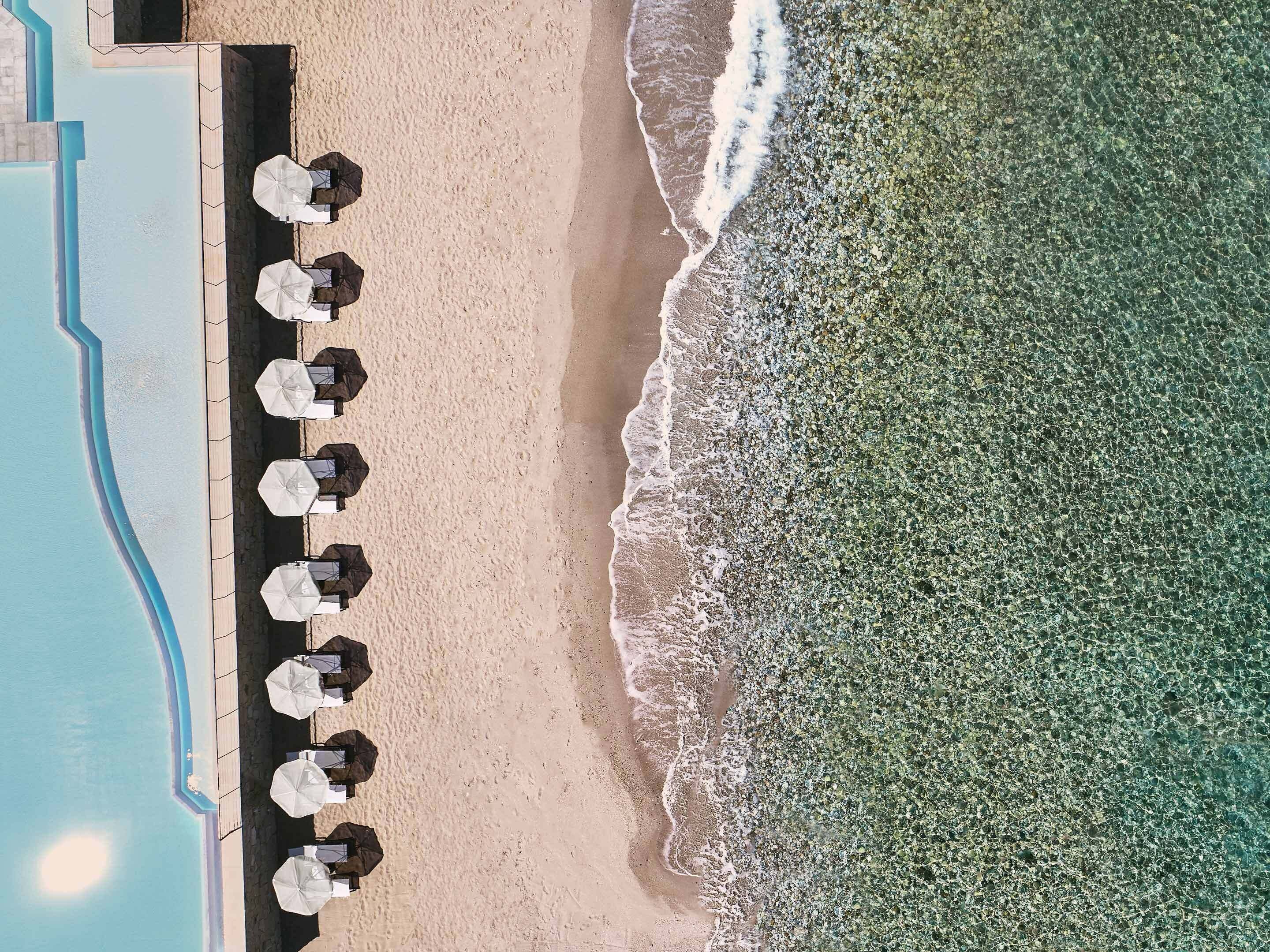 Vista aérea de una playa con sombrillas y sillas de playa dispuestas a lo largo de la orilla, junto a una piscina.