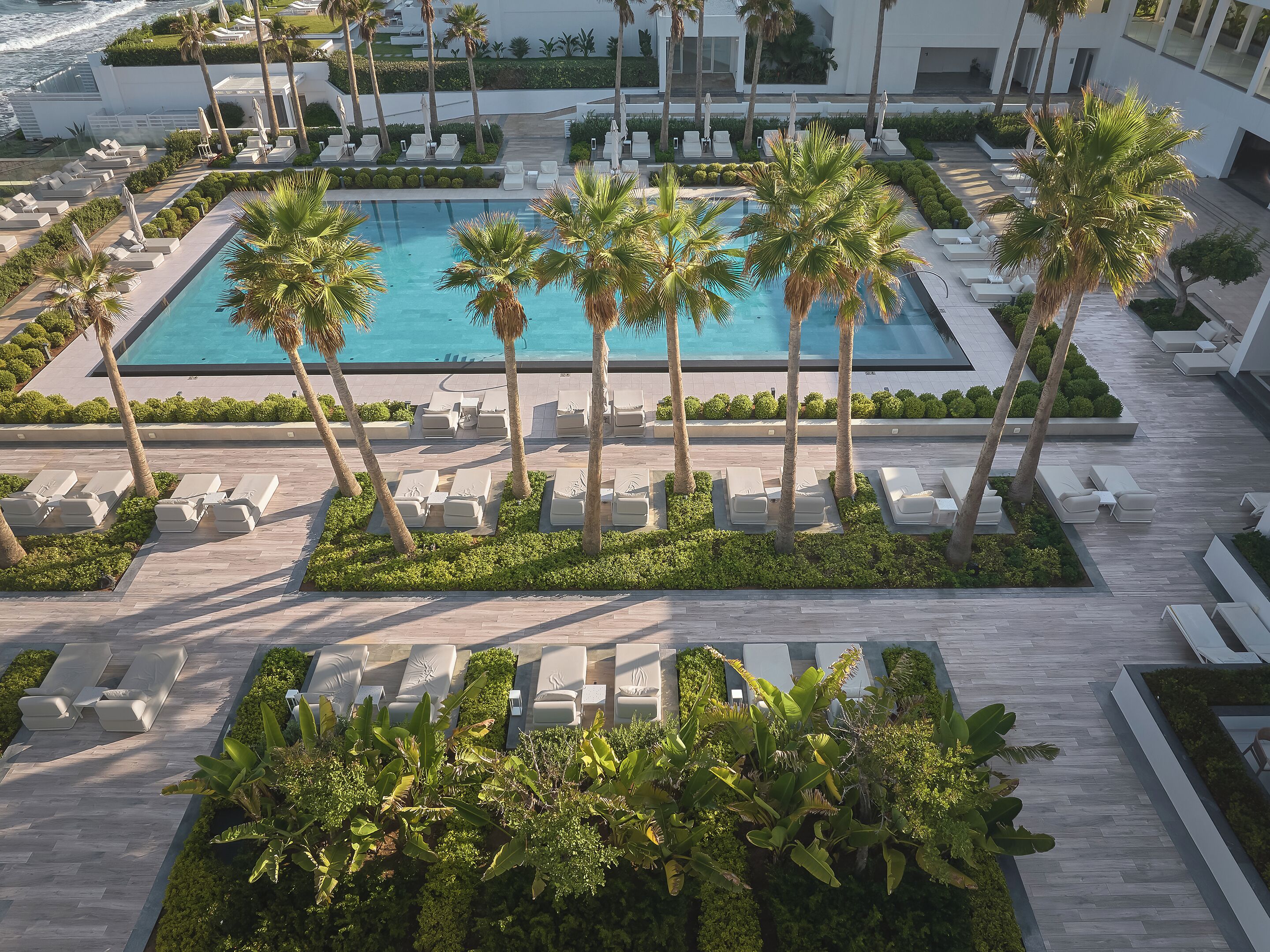 An aerial view of a resort with a central swimming pool surrounded by palm trees and sun loungers.