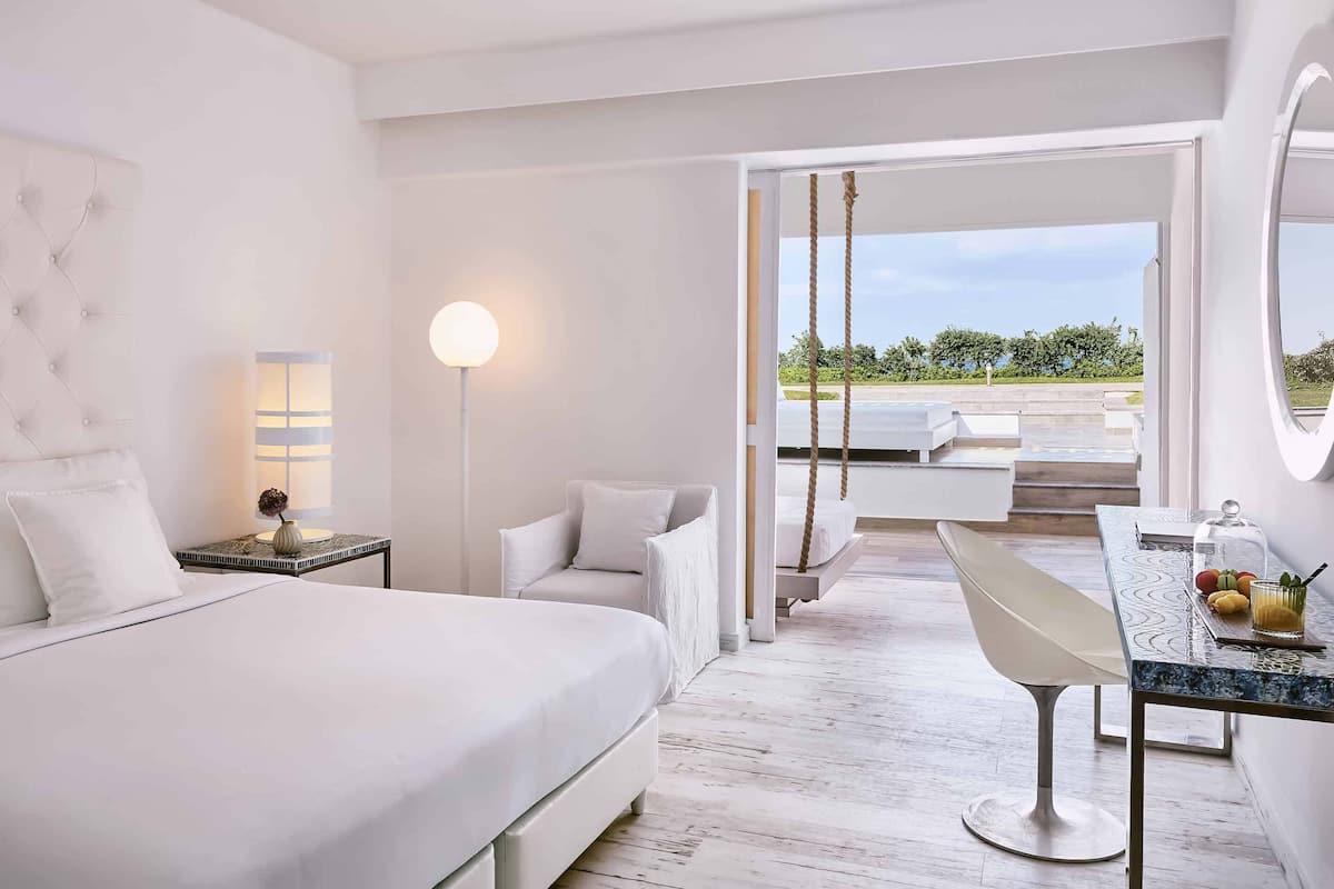A modern hotel room with a large bed, a desk with a chair, a hanging swing, and a view of the outdoors.