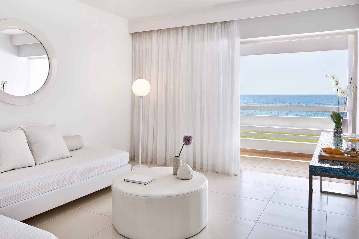 A modern hotel room with a white sofa, a round ottoman, a floor lamp, and a table with a vase and a martini glass.