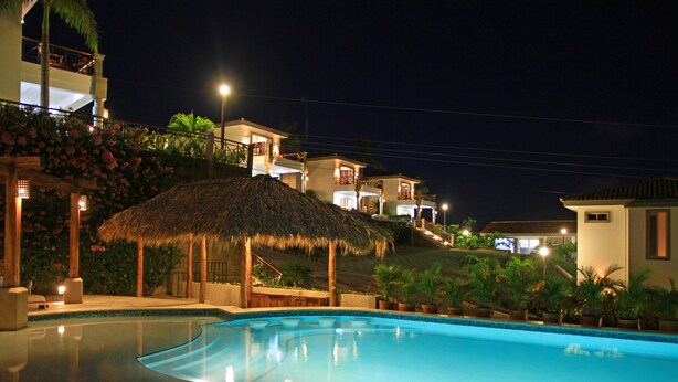 A swimming pool with a thatched roof structure, surrounded by potted plants and buildings with balconies. - Bahia del Sol Villas & Condominiums (San Juan del Sur)