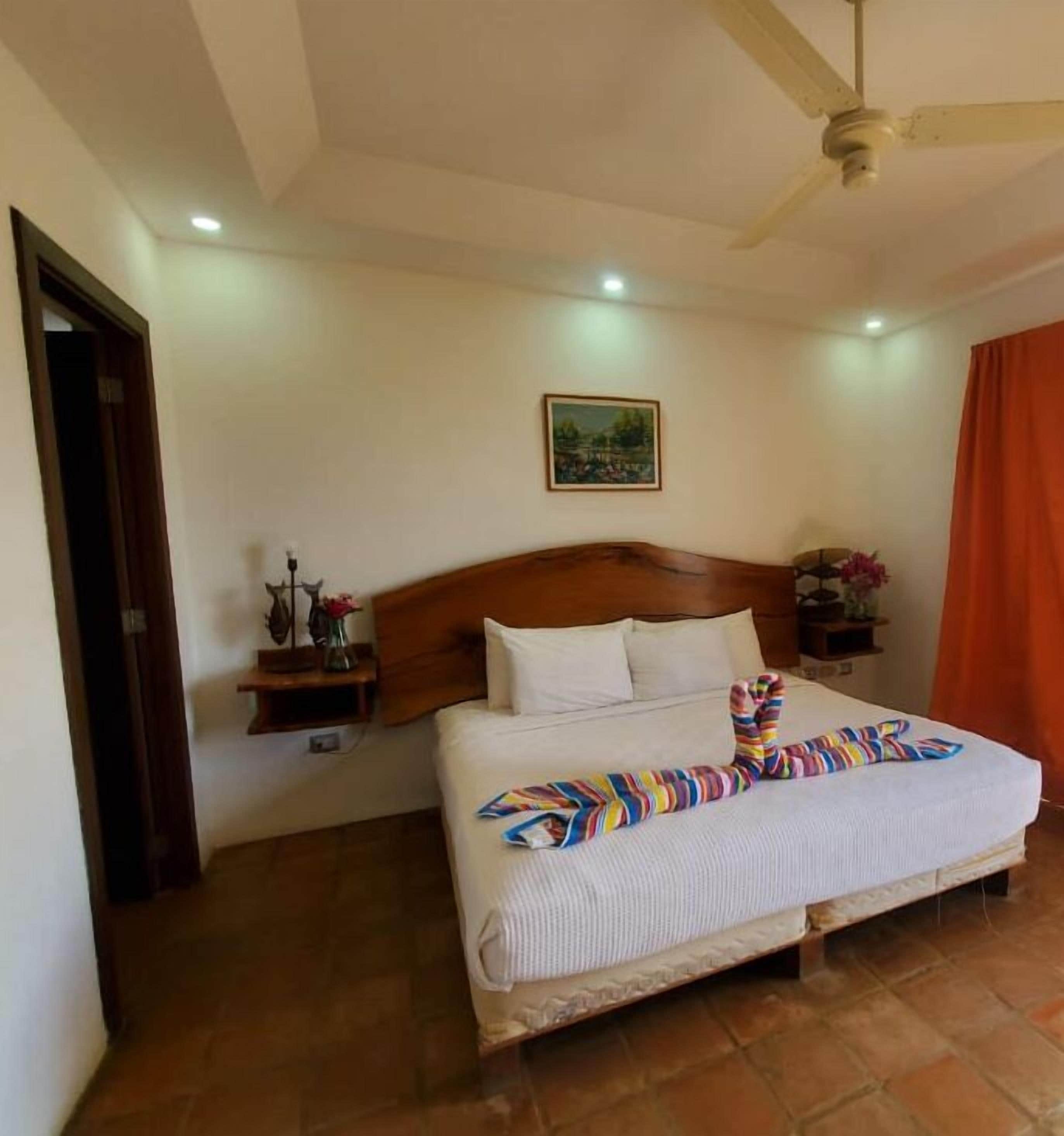 A hotel room with a large bed, a wooden headboard, a ceiling fan, and a painting on the wall.