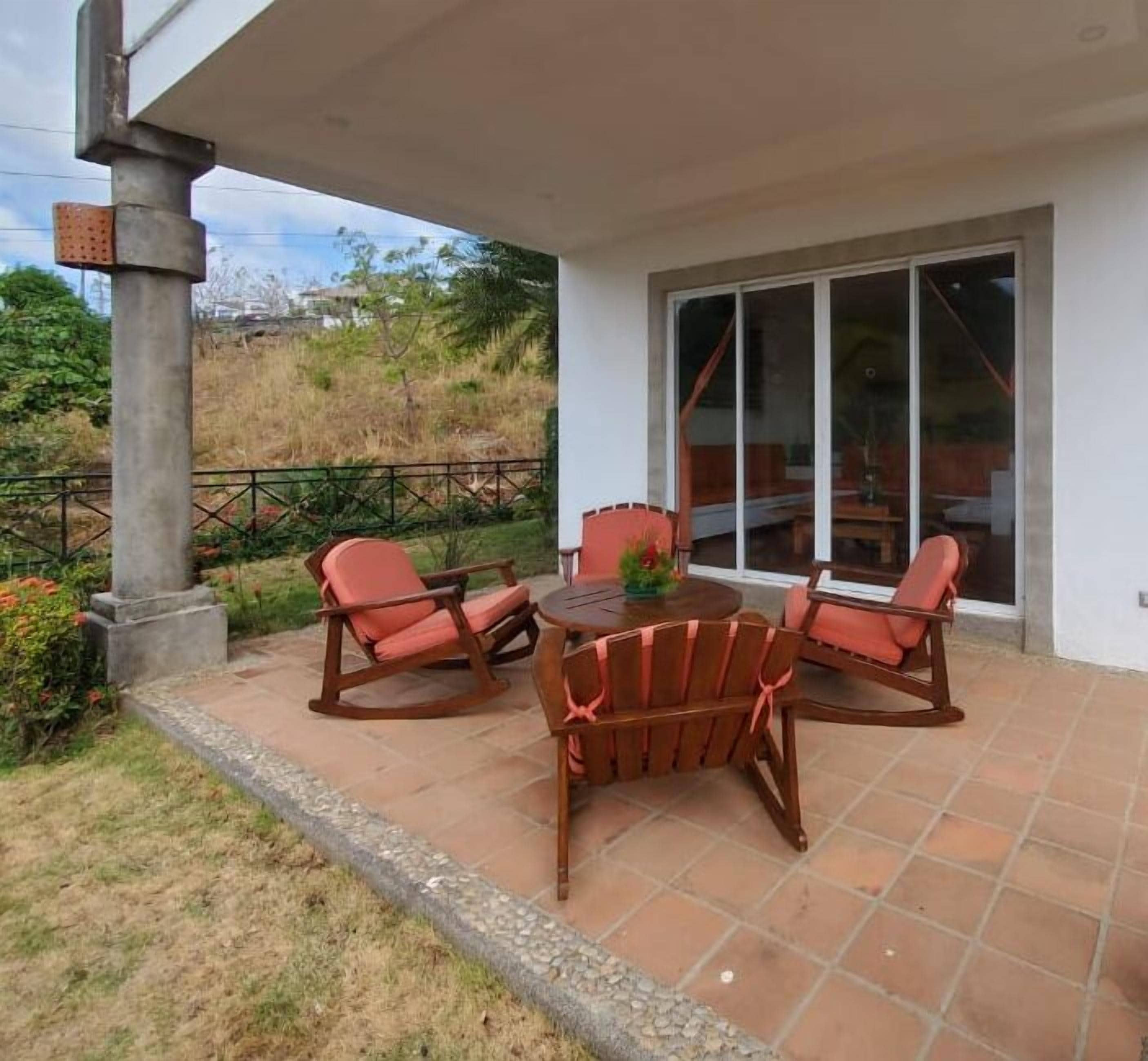 A patio area with a table and four rocking chairs, a glass door leading to a covered space, and a view of a hillside.