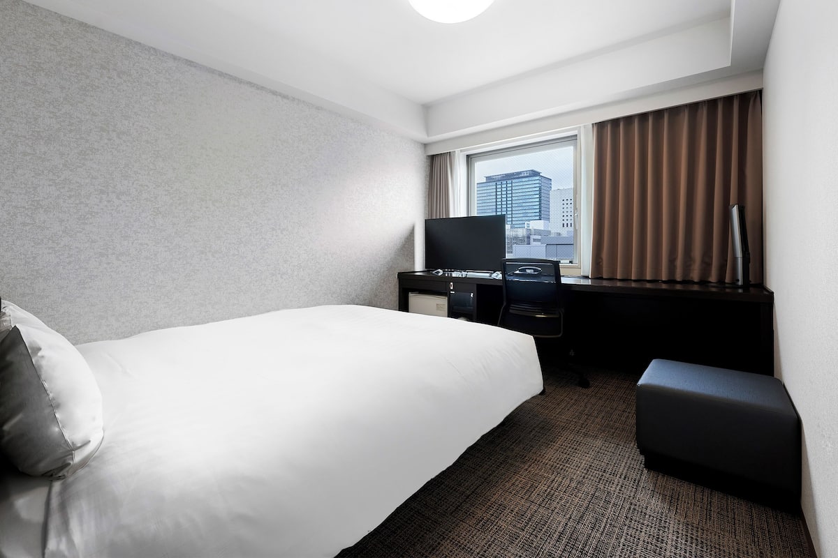 A hotel room with a bed, a desk with a TV, a chair, and a window with curtains.