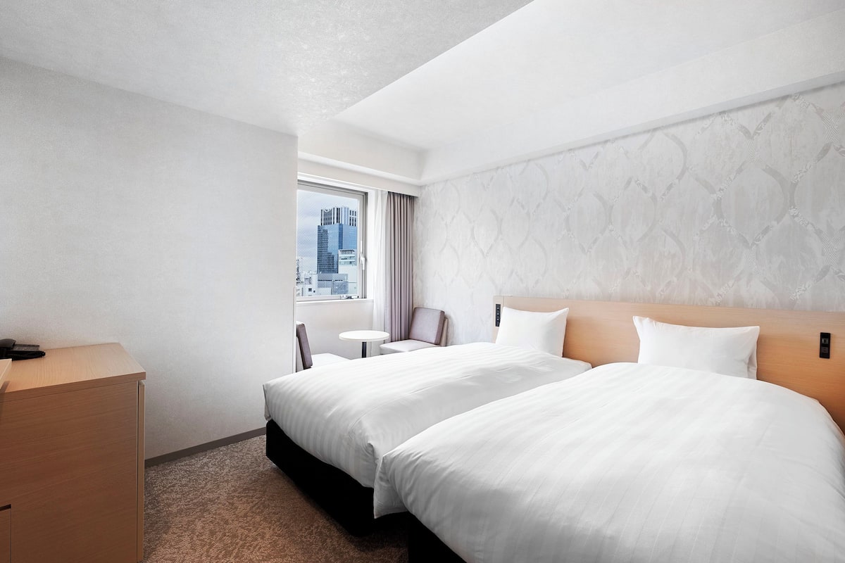 A hotel room with two beds, a desk, a chair, a small table, and a view of the cityscape through the window.