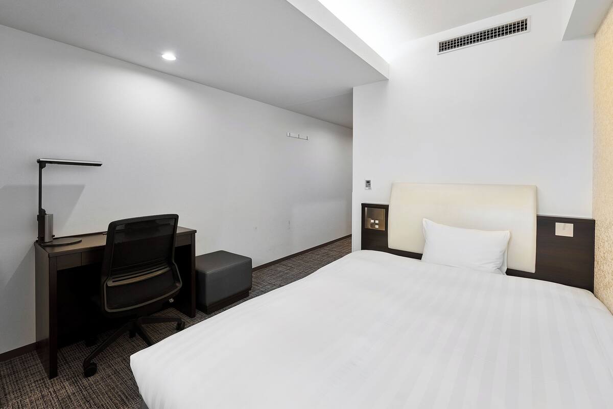 A hotel room with a bed, desk, chair, and a nightstand with a lamp.