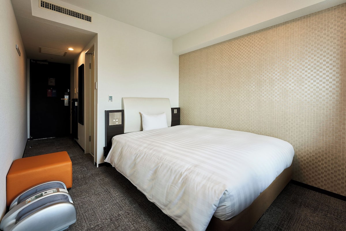 A hotel room with a large bed, a nightstand, a desk, and an orange chair.