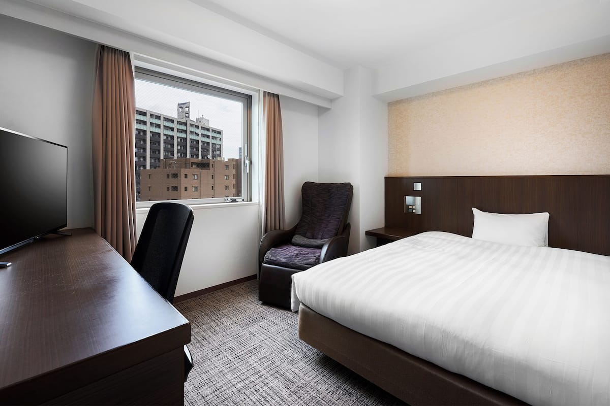 A hotel room with a bed, a desk, a chair, a TV, and a view of a building through the window.