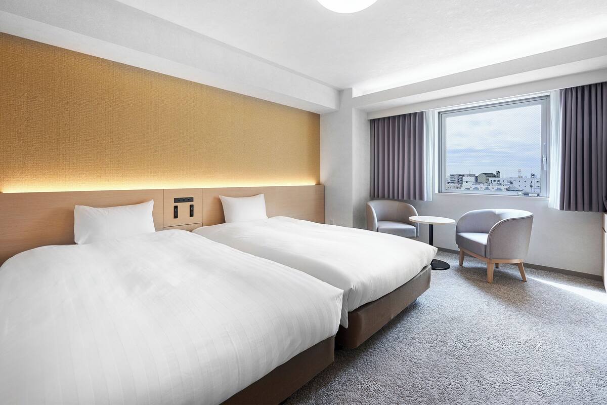 A hotel room with two beds, a small table, two chairs, a window with curtains, and a city view.