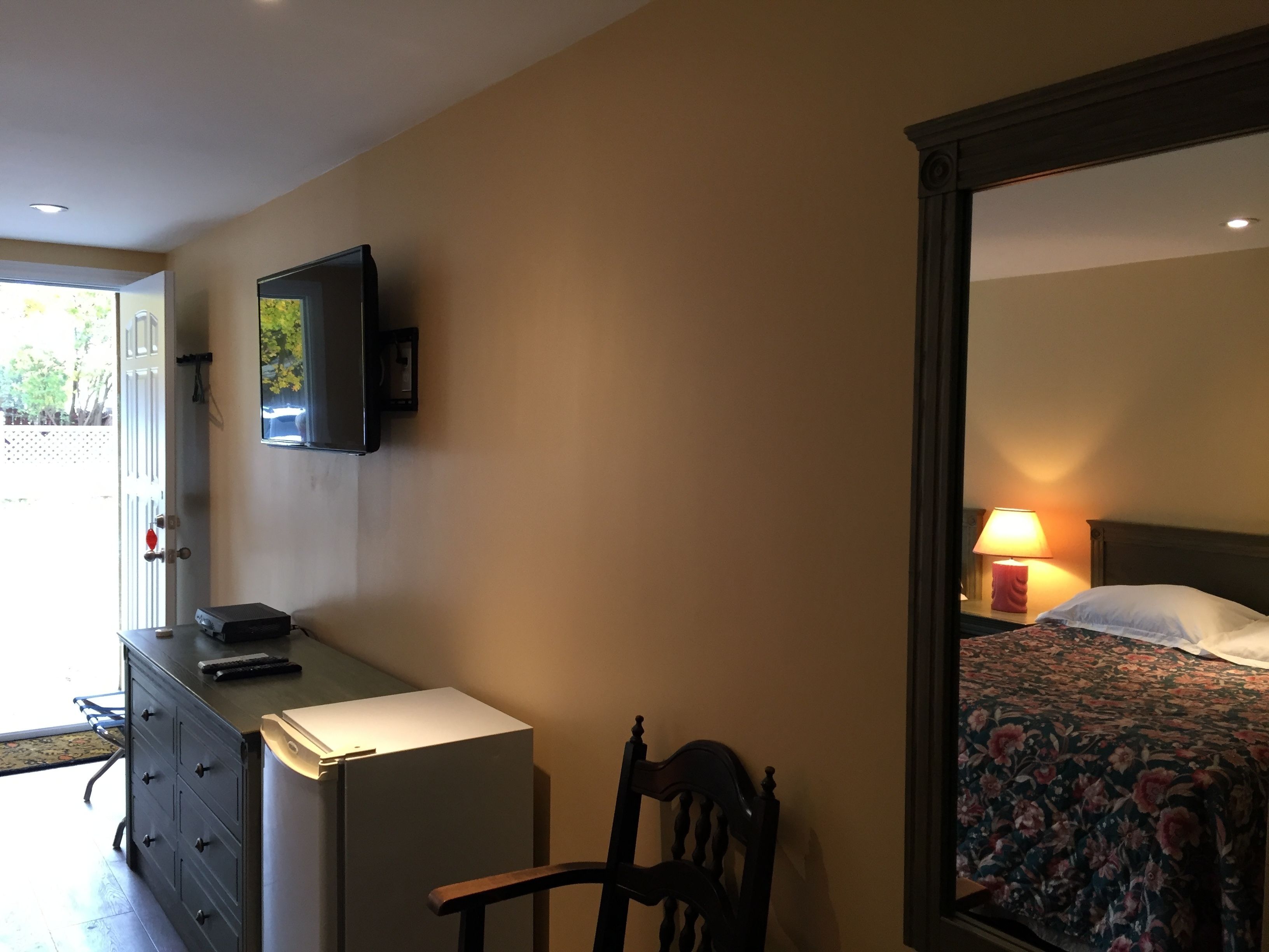 Habitación de hotel con una cama, un escritorio con computadora, una silla, un televisor montado en la pared y una puerta que da al exterior.