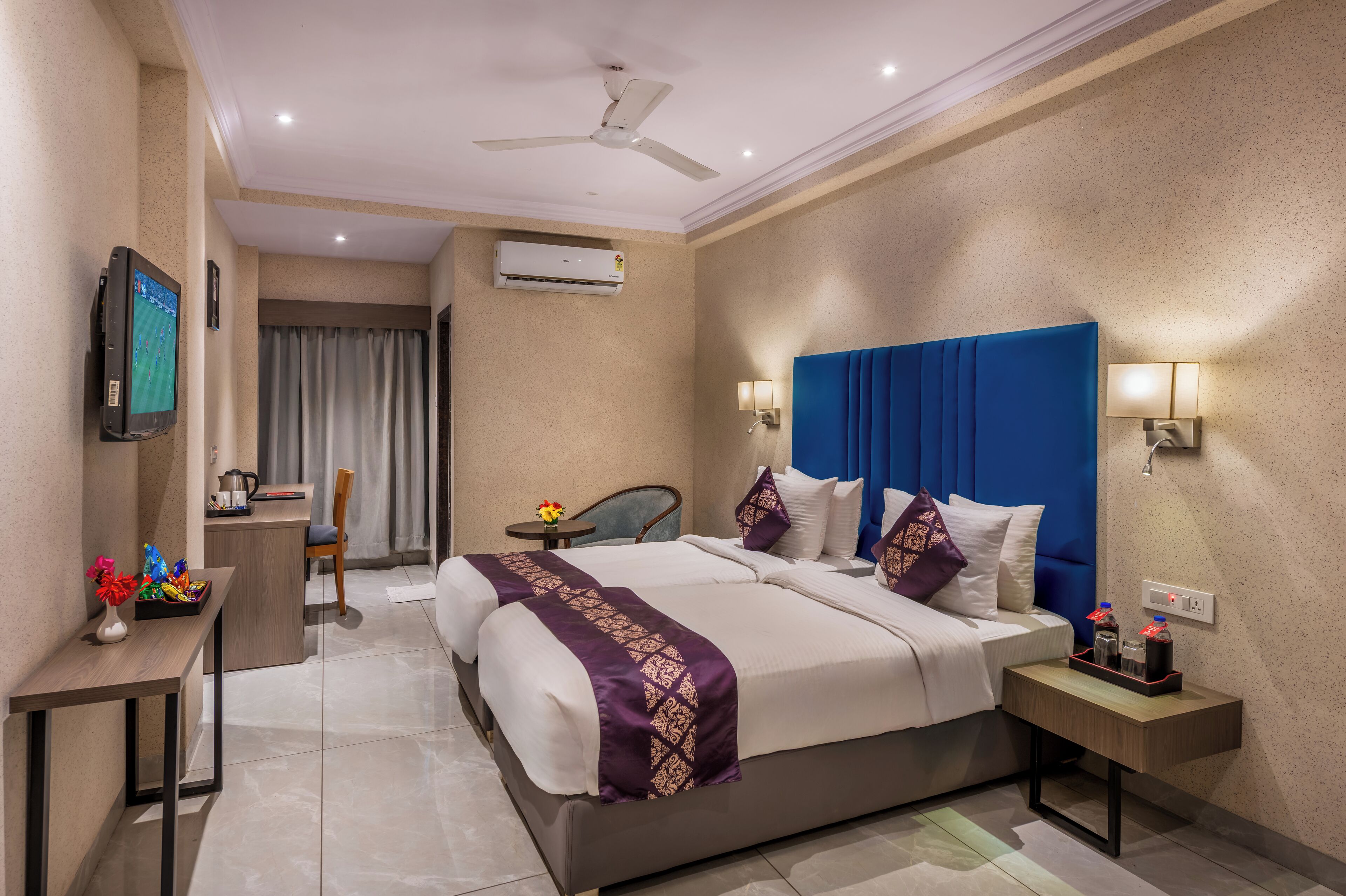 A hotel room with a large bed, a desk, a chair, a TV, an air conditioner, and a ceiling fan.