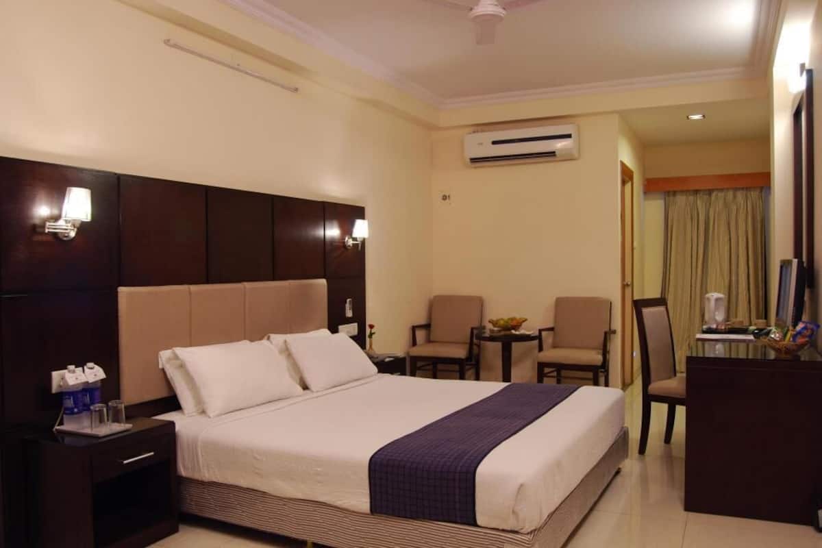 A hotel room with a large bed, bedside tables, a desk, two chairs, an air conditioning unit, and a ceiling fan.