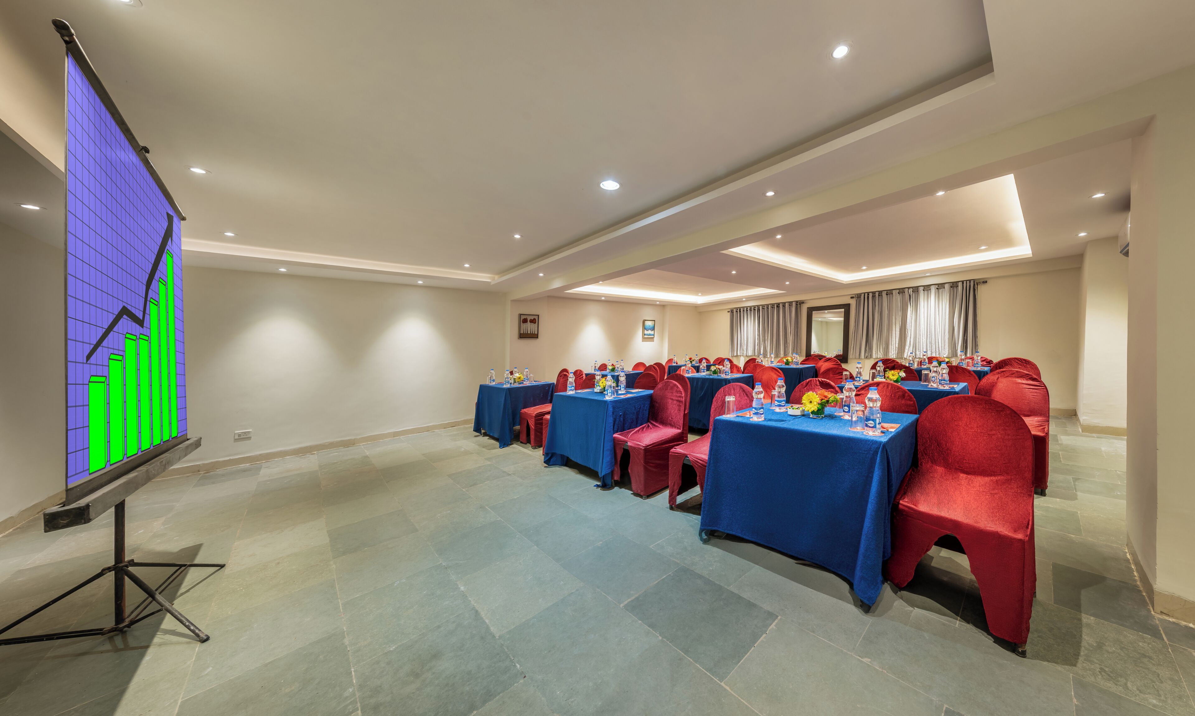Un salón de conferencias con mesas y sillas, una pantalla de proyección mostrando un gráfico y un suelo de baldosas.