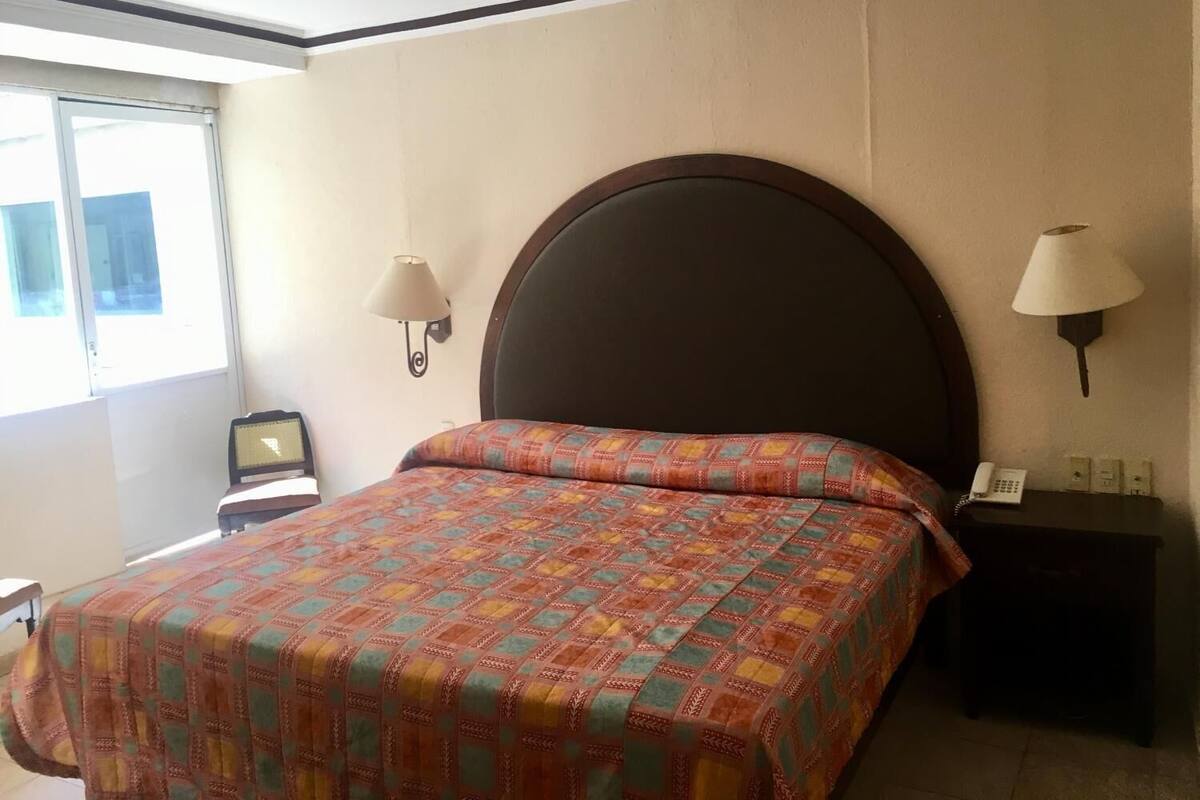 A hotel room with a large bed, two wall-mounted lamps, a small bedside table with a phone, and a window with blinds.