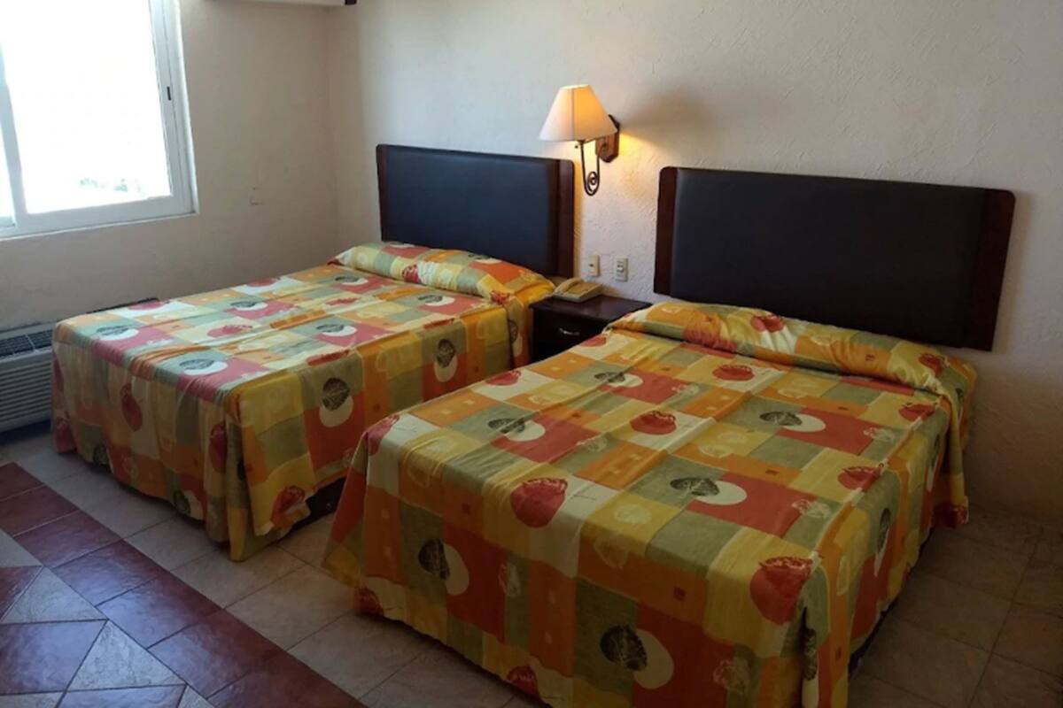 A hotel room with two beds, each with a headboard and a lamp on the nightstand.