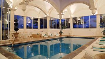 Piscina interna com cadeiras de descanso brancas e vista do céu noturno através de grandes janelas.
