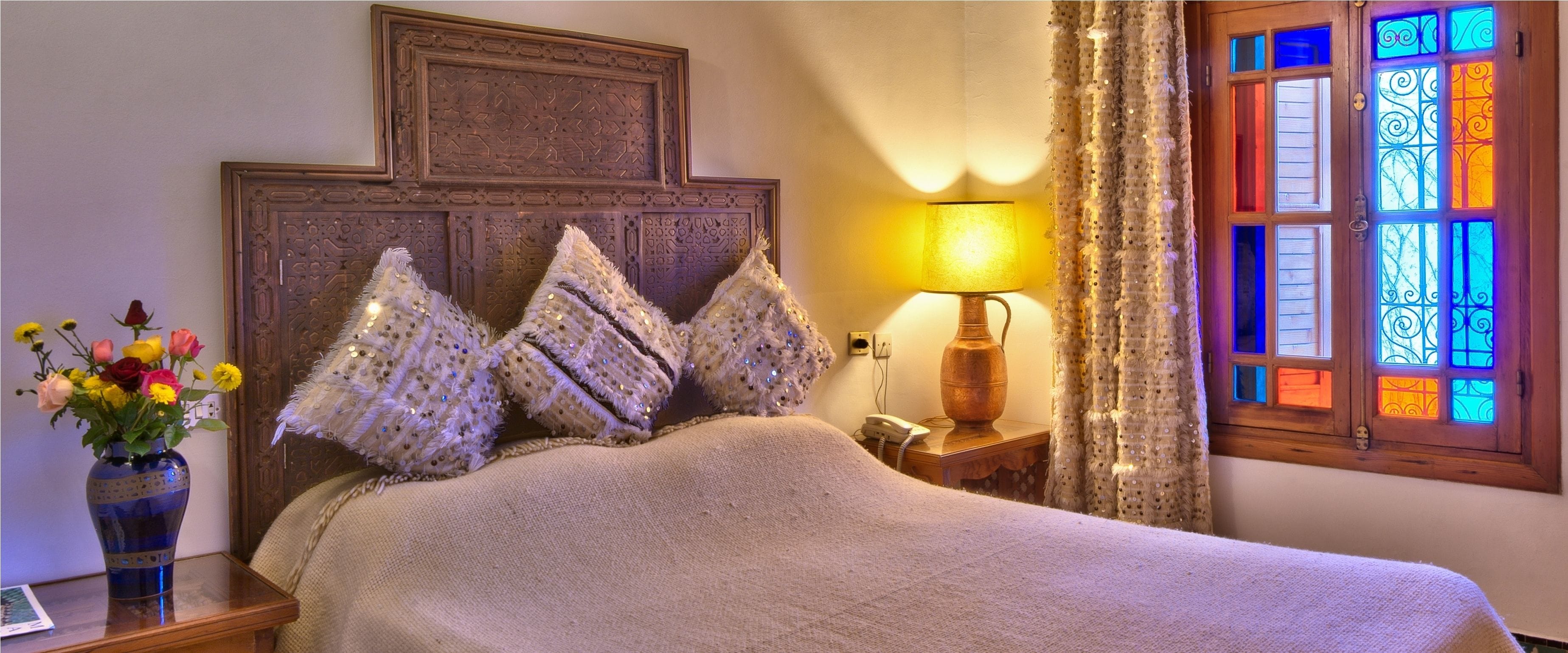 A bed with a decorative headboard, a bedside lamp, a vase with flowers, and a stained glass window.