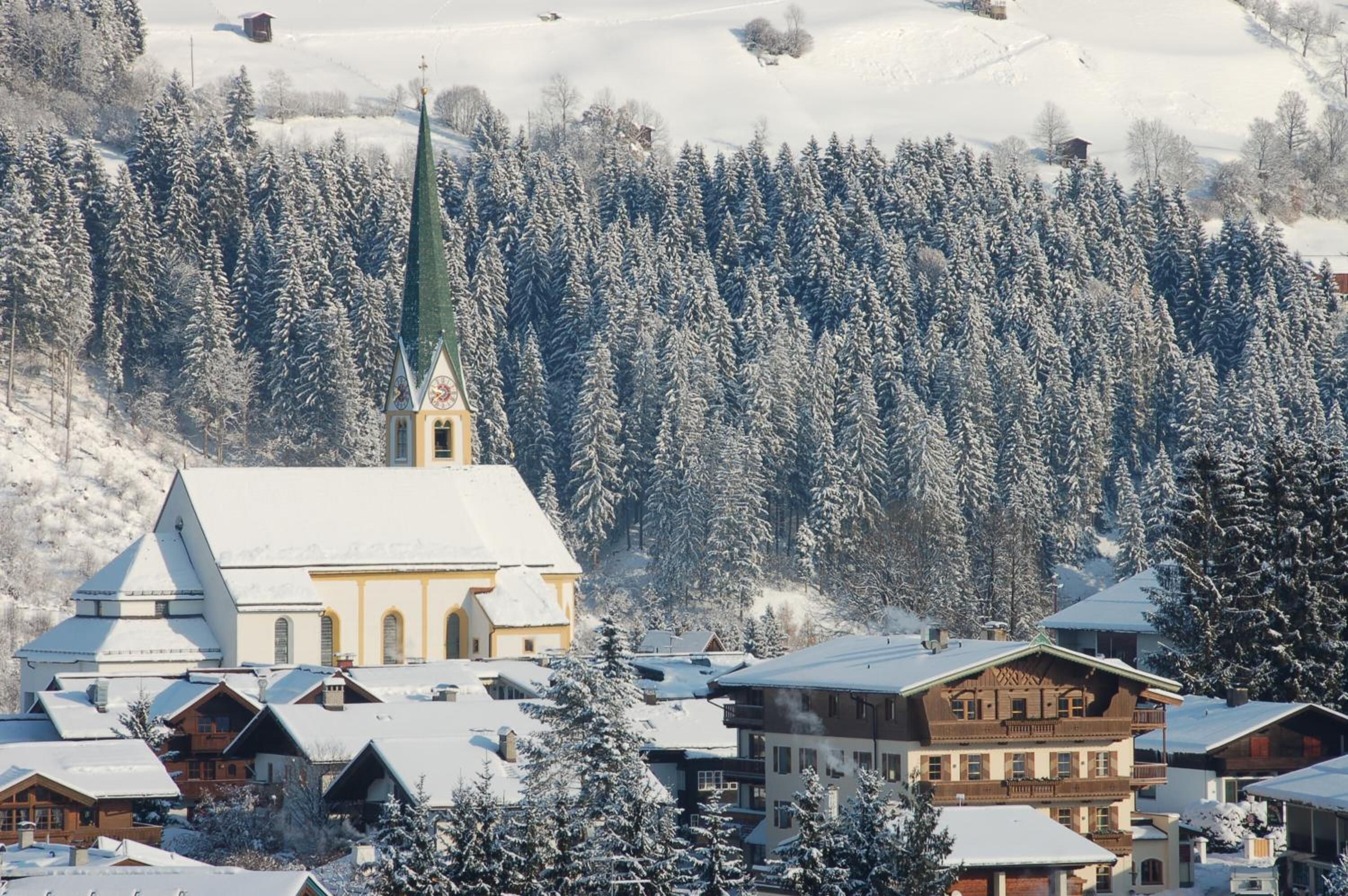 A snow-covered village with a prominent church, traditional wooden houses, and a forested mountain backdrop.
