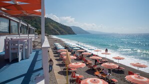 A beachside area with rows of beach chairs and umbrellas, a wooden deck, and a view of the ocean and hills in the background.